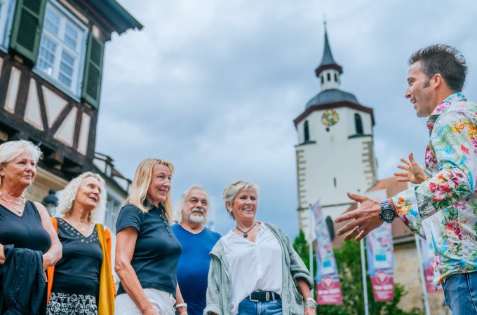 Eine Gruppe von Menschen lauscht einem Stadtf&uuml;hrer vor einem Fachwerkhaus und einem Turm mit Uhr. Der Himmel ist bew&ouml;lkt., &copy; Stadt Waldenbuch