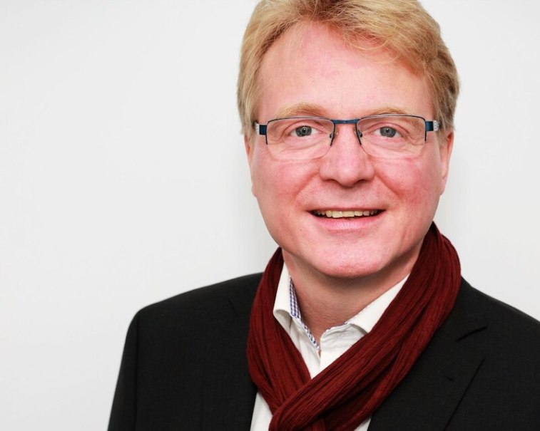 Ein Mann mit Brille und rotem Schal l&auml;chelt in die Kamera. Er tr&auml;gt einen dunklen Anzug und steht vor einem wei&szlig;en Hintergrund., &copy; Sch&ouml;nblick gemeinn&uuml;tzige GmbH