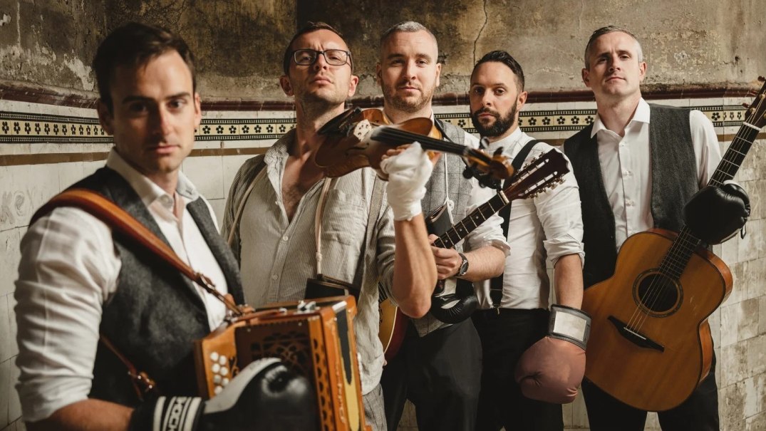 F&uuml;nf M&auml;nner in hellen Hemden und Westen mit Musikinstrumenten, hintereinander in einer Reihe in einer Zimmerecke stehend, &copy; The Rapparees