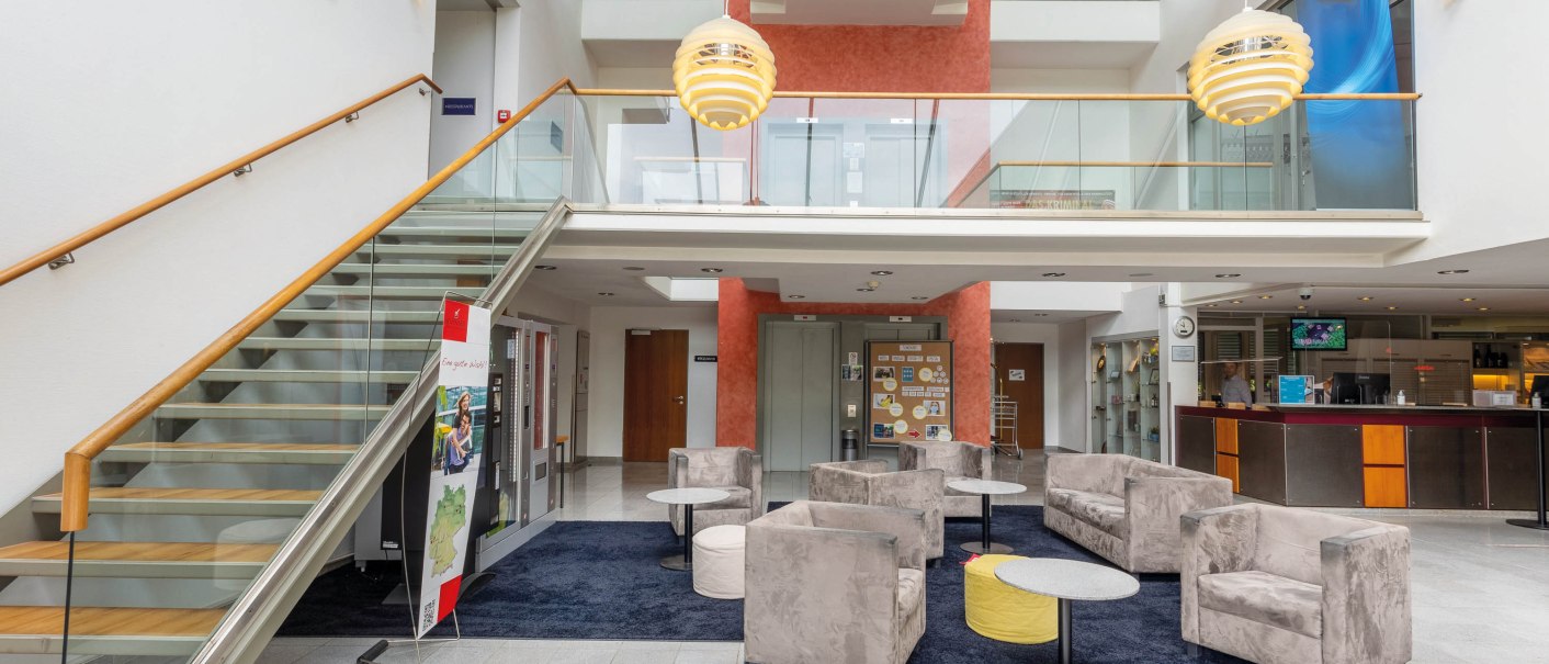 Moderne Lobby mit Sitzbereich, grauen Sesseln, Treppe mit Glasgeländer und Rezeption. Helle Beleuchtung und offene Architektur., © Seminaris Hotel Bad Boll Moderne Lobby mit Sitzbereich, grauen Sesseln, Treppe mit Glasgeländer und Rezeption. Helle Beleuchtung und offene Architektur., © Seminaris Hotel Bad Boll