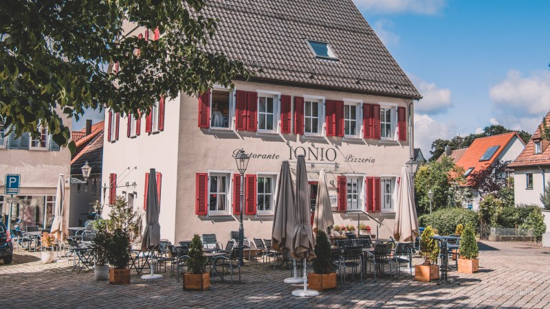 Ein Restaurant mit roten Fensterläden und Außensitzbereich auf einem gepflasterten Platz, umgeben von Bäumen und blauem Himmel., © Stadt Welzheim