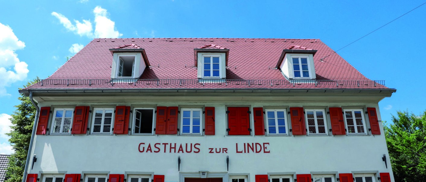 Das Gasthaus zur Linde hat rote Fensterläden und ein rotes Ziegeldach. Der Himmel ist blau mit wenigen Wolken., © Fei Shiyu Das Gasthaus zur Linde hat rote Fensterläden und ein rotes Ziegeldach. Der Himmel ist blau mit wenigen Wolken., © Fei Shiyu