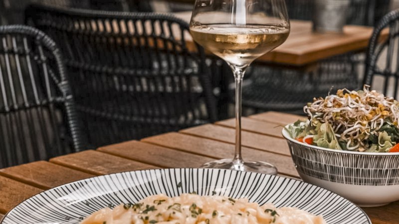 Ein Glas Weißwein, ein Teller Pasta und ein Salat auf einem Holztisch in einem Restaurant mit geflochtenen Stühlen., © SMG, itchyfeet-travel.de