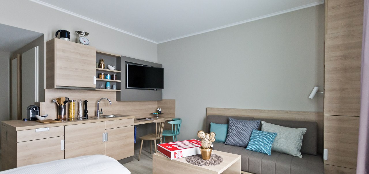 Moderne Einzimmerwohnung mit Küchenzeile, Sofa, Fernseher und Esstisch. Helle Holzmöbel und neutrale Farben dominieren den Raum., © Arthotel Ana / elaya