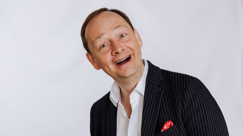 Ein Mann in gestreiftem Anzug mit rotem Einstecktuch lächelt vor weißem Hintergrund., © Stadthalle Leonberg