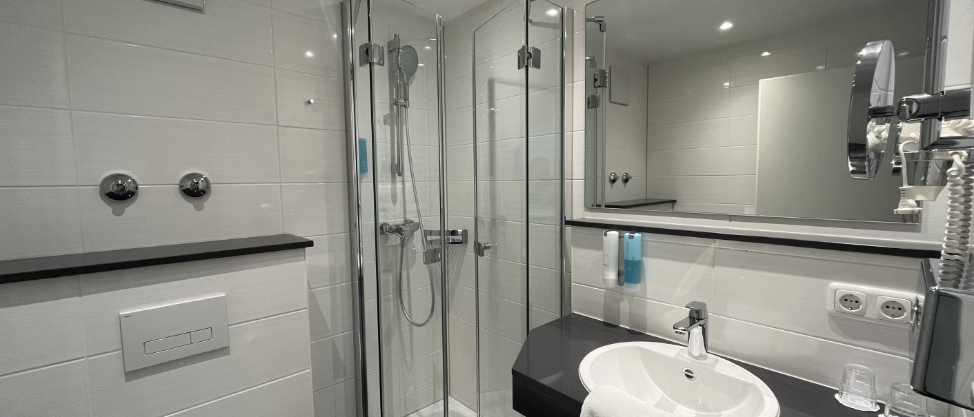 Modernes Badezimmer mit Glasdusche, Waschbecken, großem Spiegel und weißen Fliesen. Handtuch und Pflegeprodukte sind vorhanden., © TOMAS