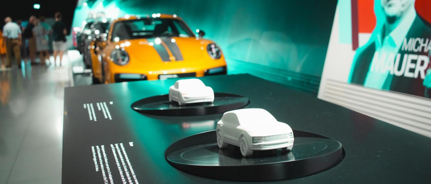 Modellautos auf einem Tisch im Porsche Museum, im Hintergrund ein orangefarbener Porsche und Besucher. Rechts ein Plakat mit dem Namen Michael Mauer., © Stuttgart Marketing GmbH, Sarah Schmid