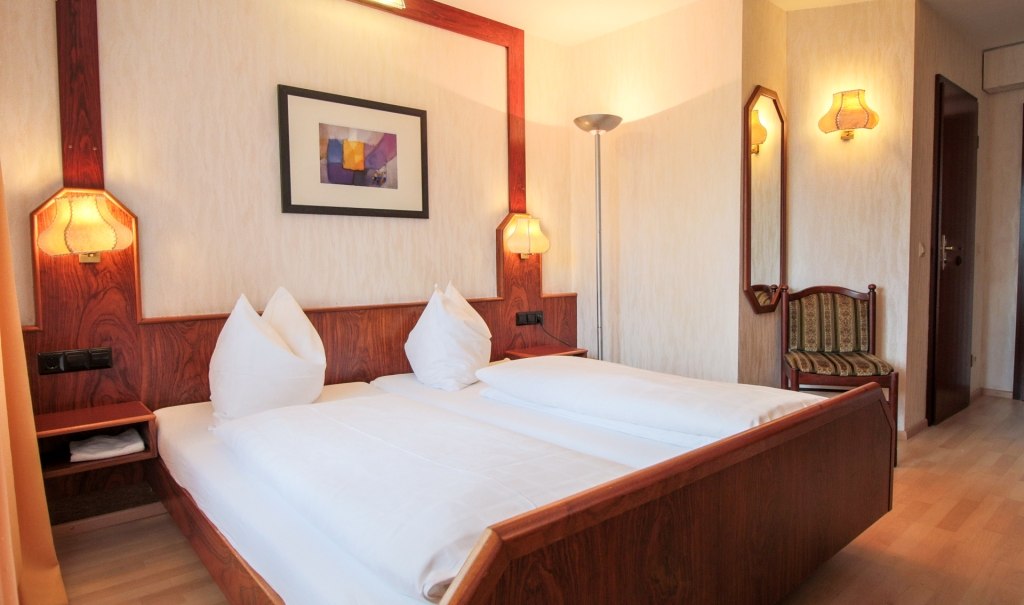 Ein komfortables Doppelzimmer mit Holzbetten, zwei Nachttischlampen, einem Kunstwerk an der Wand und einem Stuhl in der Ecke., © Ulli Kellenbenz Ein komfortables Doppelzimmer mit Holzbetten, zwei Nachttischlampen, einem Kunstwerk an der Wand und einem Stuhl in der Ecke., © Ulli Kellenbenz