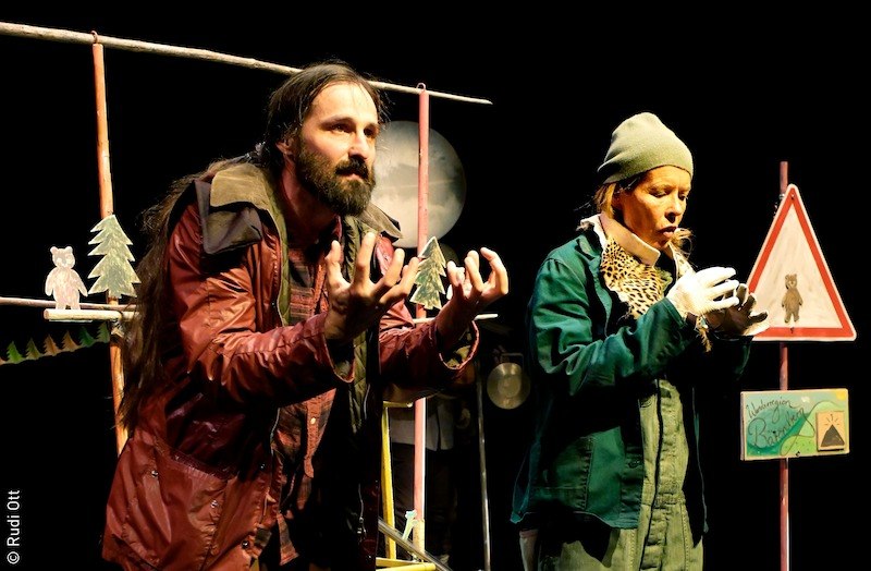Zwei Schauspieler auf der Bühne in einem Theaterstück. Sie tragen Outdoor-Kleidung, im Hintergrund sind Waldkulisse und Bärenschilder zu sehen., © Stadt Kornwestheim Kulturmanagement