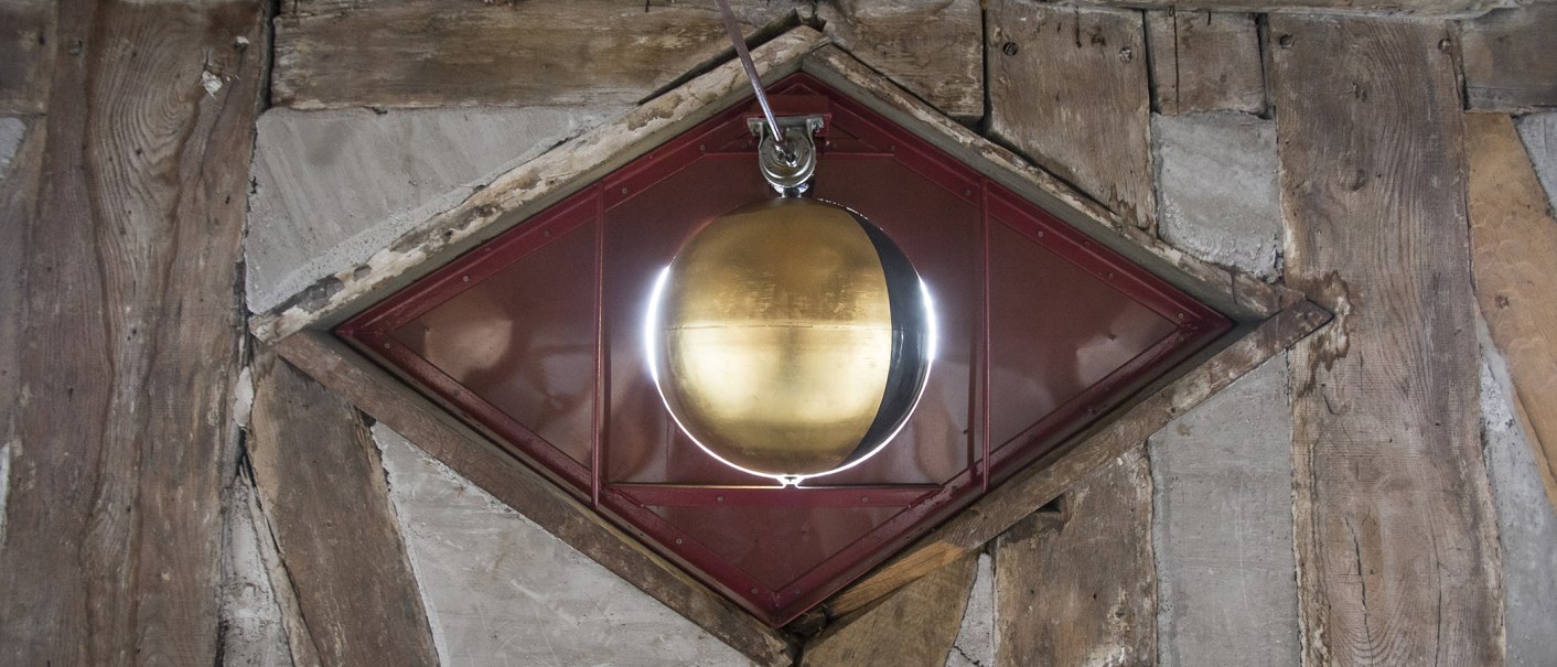 Ein goldenes Pendel hängt in einem rautenförmigen Holzrahmen. Der Hintergrund besteht aus alten Holzbalken und Mauerwerk., © Torsten Wenzler