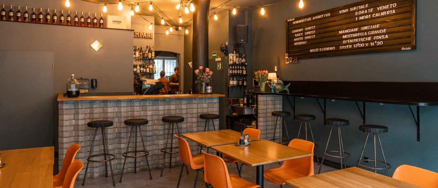 Gemütliche Bar mit Holztischen, Barhockern und einer Theke. Lichterketten und ein Menübrett schmücken die Wand., © SMG, Sarah Schmid