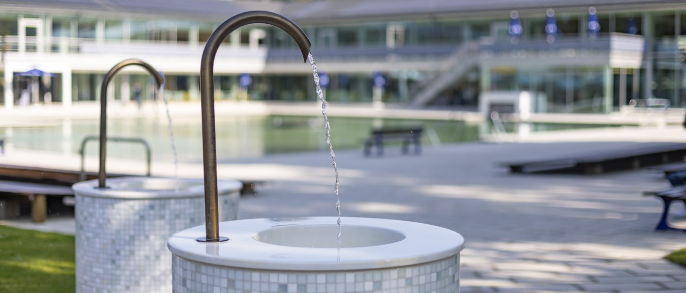 Ein Trinkbrunnen im Mineralbad Berg mit fließendem Wasser im Vordergrund, im Hintergrund ein modernes Gebäude und ein Pool., © Stuttgarter Bäder