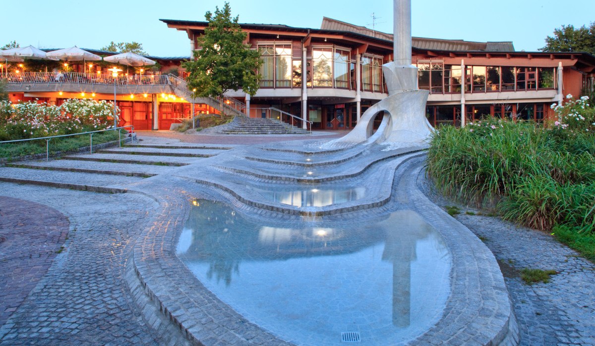 Moderner Brunnen mit geschwungenen Formen und Wasserbecken vor einem beleuchteten Gebäude mit Terrasse und Pflanzen., © Waiblingen - Stuttgart-Marketing GmbH Moderner Brunnen mit geschwungenen Formen und Wasserbecken vor einem beleuchteten Gebäude mit Terrasse und Pflanzen., © Waiblingen - Stuttgart-Marketing GmbH