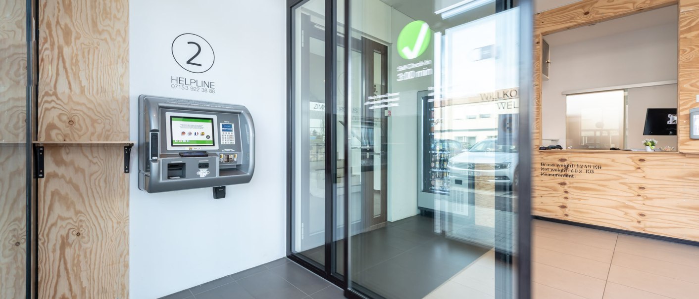 Moderner Eingangsbereich mit Selbstbedienungsterminal an der Wand, Glastür und Empfangstresen aus Holz., © a2 HOTELS