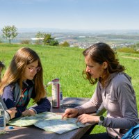 Drei Personen sitzen an einem Tisch im Freien und betrachten eine Karte. Im Hintergrund ist eine weite Landschaft mit Feldern und Bäumen zu sehen., © Stadtverwaltung Winnenden