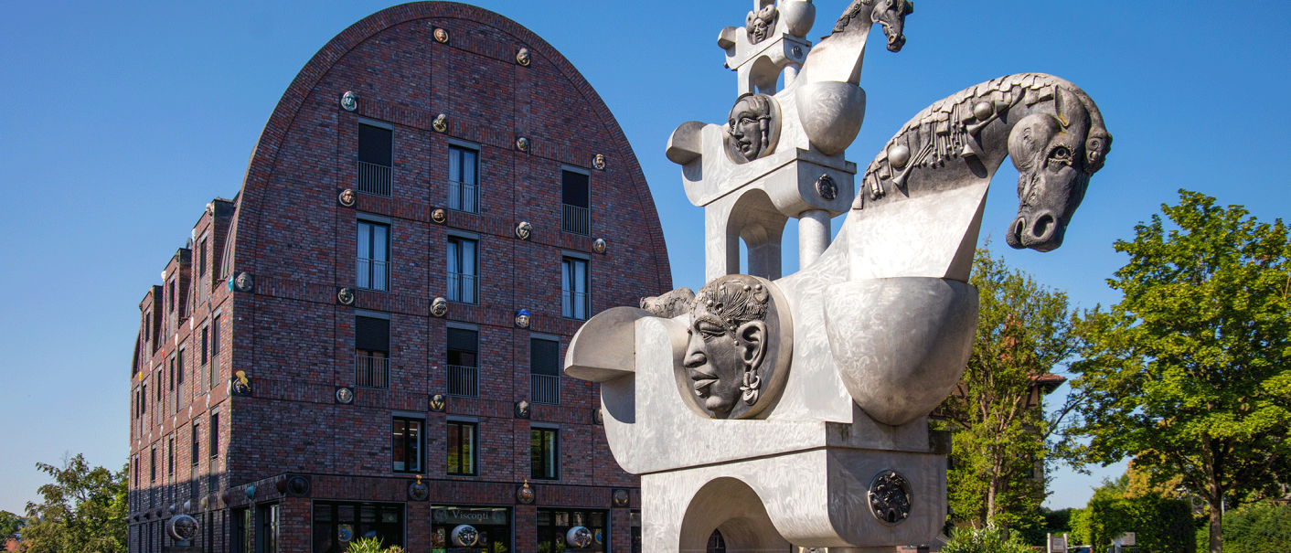 Moderne Skulptur mit Pferde- und Menschenköpfen vor einem Backsteingebäude mit rundem Dach, umgeben von Bäumen und Pflanzen., © Stuttgart-Marketing GmbH, Achim Mende