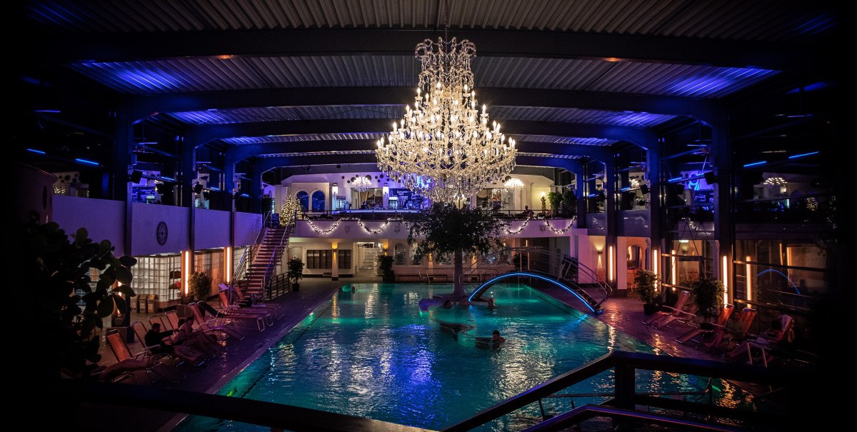 Luxuriöses Spa mit großem Kronleuchter über einem beleuchteten Pool, umgeben von Liegestühlen und Pflanzen. Elegante Atmosphäre mit blauer Beleuchtung., © AlbThermen Bad Urach