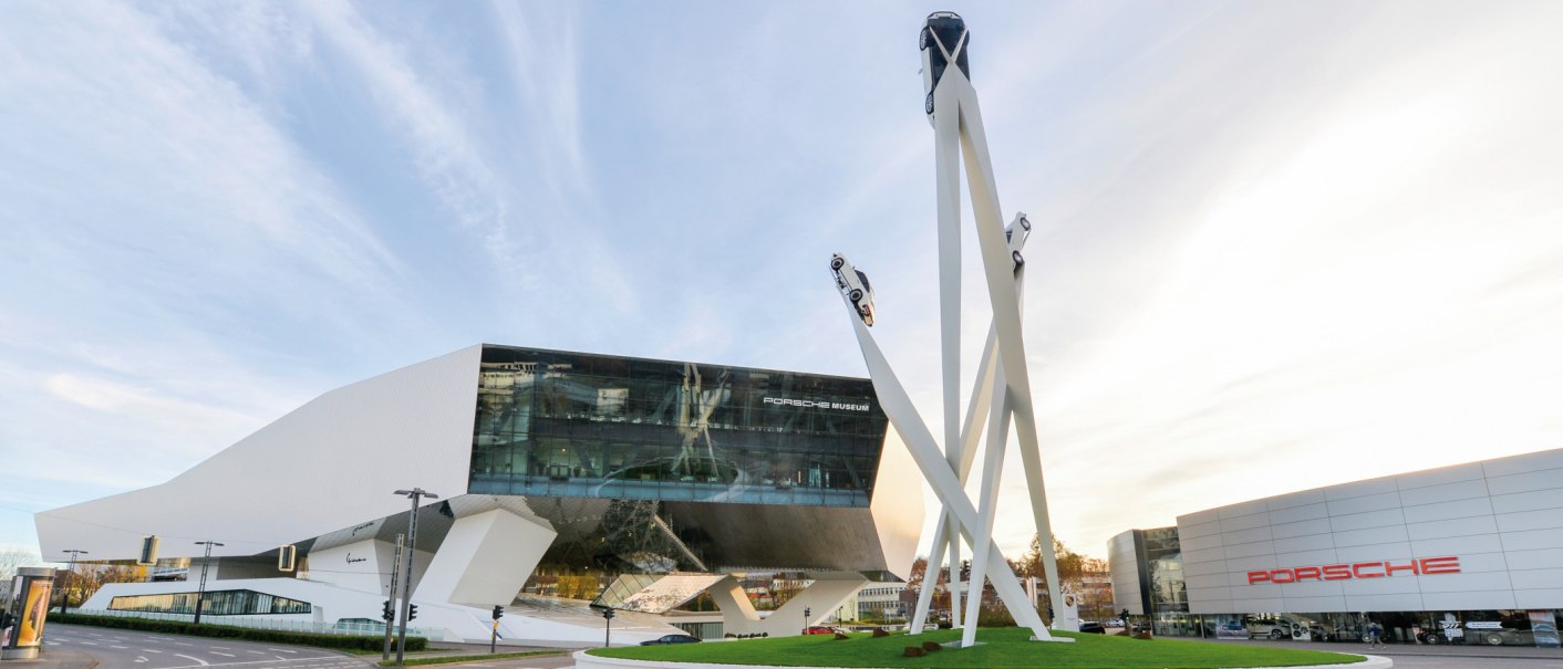Das Porsche Museum mit seiner modernen Architektur und einer Skulptur von drei Autos auf hohen Säulen vor einem blauen Himmel., © Porsche AG Sabine Braun