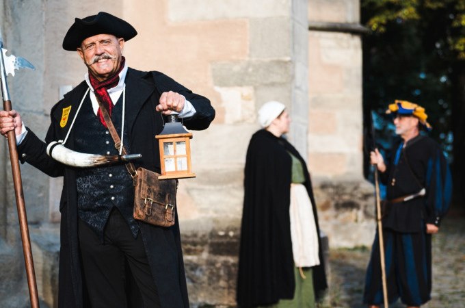 Person in historischer Kleidung mit Laterne und Hellebarde, zwei weitere Personen im Hintergrund vor einem alten Gebäude., © Stadt Nürtingen
