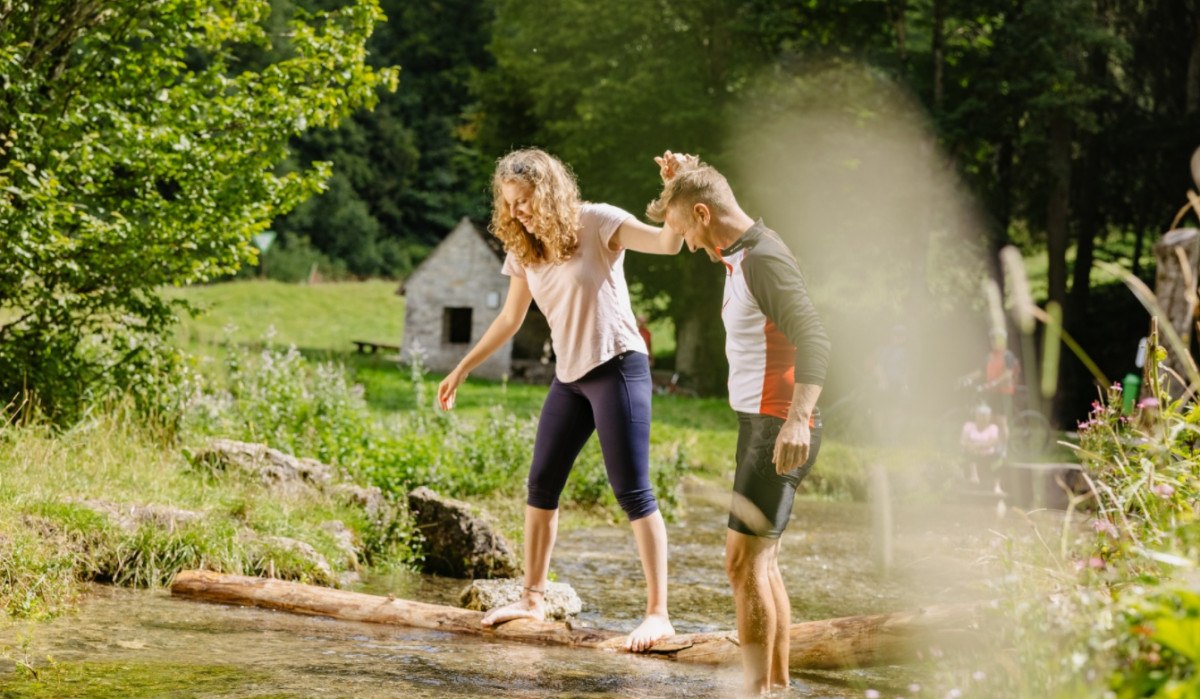 Zwei Personen balancieren auf einem Baumstamm über einen Bach. Im Hintergrund sind Bäume und eine kleine Hütte zu sehen., © Landkreis Göppingen Zwei Personen balancieren auf einem Baumstamm über einen Bach. Im Hintergrund sind Bäume und eine kleine Hütte zu sehen., © Landkreis Göppingen