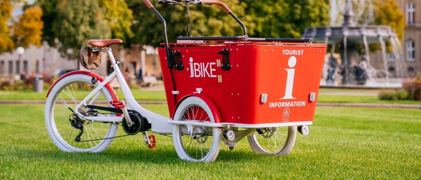 Ein rotes Lastenfahrrad mit der Aufschrift 'I-Bike' steht auf einer Wiese vor einem Brunnen und einem Gebäude mit Kuppel., © Stuttgart-Marketing GmbH, Thomas Niedermüller Ein rotes Lastenfahrrad mit der Aufschrift 'I-Bike' steht auf einer Wiese vor einem Brunnen und einem Gebäude mit Kuppel., © Stuttgart-Marketing GmbH, Thomas Niedermüller