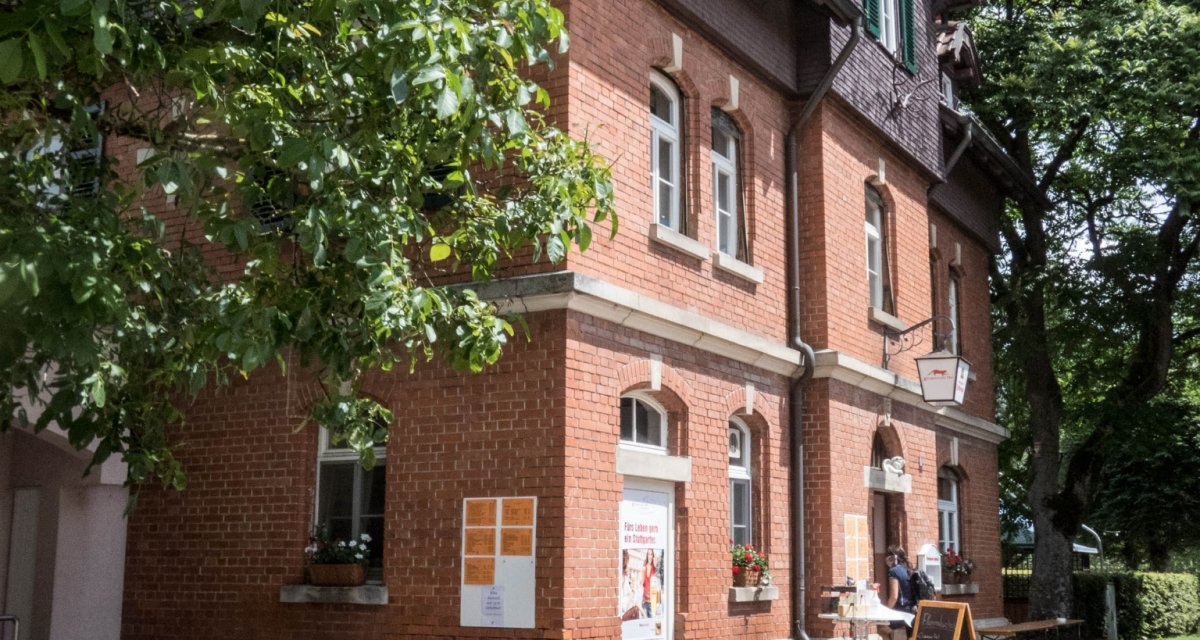 Ein Backsteingebäude mit grünen Fensterläden, umgeben von Bäumen. Vor dem Gebäude stehen Bierbänke und eine Tafel auf dem Gehweg., © Natur.Nah. Schönbuch & Heckengäu Ein Backsteingebäude mit grünen Fensterläden, umgeben von Bäumen. Vor dem Gebäude stehen Bierbänke und eine Tafel auf dem Gehweg., © Natur.Nah. Schönbuch & Heckengäu
