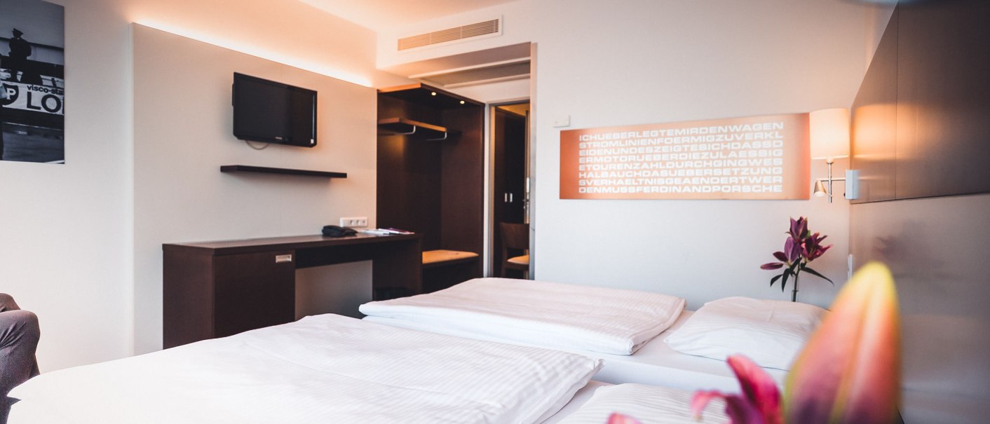 Modernes Hotelzimmer mit Doppelbett, Wand-TV, Schreibtisch und Blumen. Dezente Beleuchtung und stilvolle Einrichtung schaffen eine gemütliche Atmosphäre., © Abacco Hotels GmbH Modernes Hotelzimmer mit Doppelbett, Wand-TV, Schreibtisch und Blumen. Dezente Beleuchtung und stilvolle Einrichtung schaffen eine gemütliche Atmosphäre., © Abacco Hotels GmbH