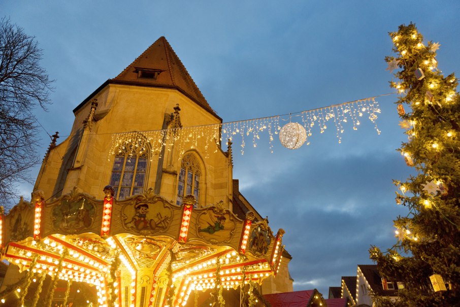 Eine festlich beleuchtete Stadtkirche mit Lichterketten, einem Karussell und einem geschmückten Weihnachtsbaum im Vordergrund., © Stadt Nürtingen
