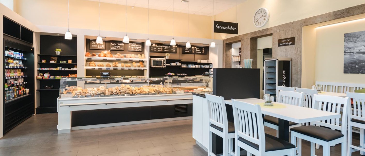 Moderne Bäckerei mit Verkaufstheke voller Backwaren, weißen Tischen und Stühlen. Helle Beleuchtung und Regale mit Snacks im Hintergrund., © Bäckerei Veit, Dr. Michael Immendörfer