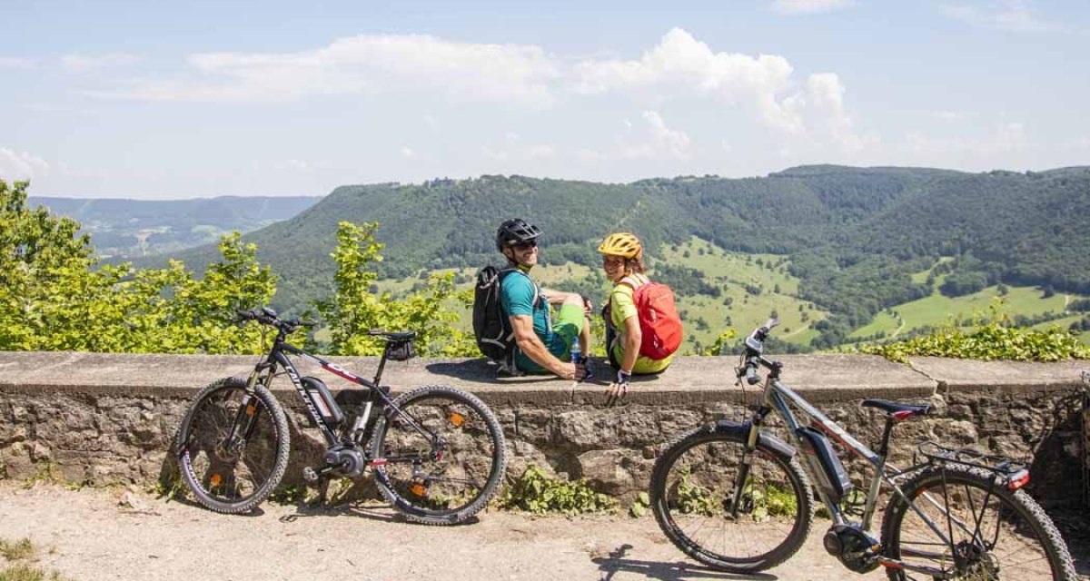 Zwei Radfahrer sitzen auf einer Mauer und genie&szlig;en den Ausblick auf die gr&uuml;ne H&uuml;gellandschaft. Zwei Fahrr&auml;der stehen daneben., &copy; Schw&auml;bische Alb Tourismusverband e.V.