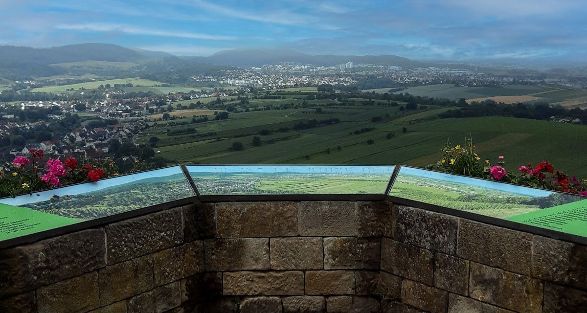 Aussichtspunkt mit Panoramatafeln, Blick auf Felder und eine Stadt in der Ferne. Blumen schmücken den Rand, blauer Himmel im Hintergrund., © Remstal Tourismus e.V. Aussichtspunkt mit Panoramatafeln, Blick auf Felder und eine Stadt in der Ferne. Blumen schmücken den Rand, blauer Himmel im Hintergrund., © Remstal Tourismus e.V.
