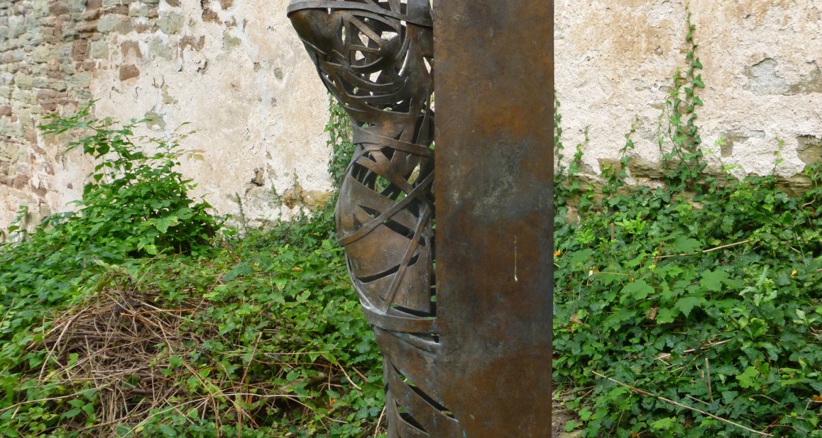 Metallskulptur eines Menschen, der hinter einer Wand hervorschaut, vor einer alten Steinmauer und umgeben von Pflanzen., © Natur.Nah. Schönbuch & Heckengäu Metallskulptur eines Menschen, der hinter einer Wand hervorschaut, vor einer alten Steinmauer und umgeben von Pflanzen., © Natur.Nah. Schönbuch & Heckengäu