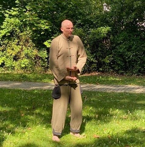 Ein Mann in traditioneller Qigong-Kleidung steht barfu&szlig; auf einer Wiese und f&uuml;hrt eine &Uuml;bung durch. Im Hintergrund sind B&auml;ume und B&uuml;sche zu sehen., &copy; Daniel Steinbauer