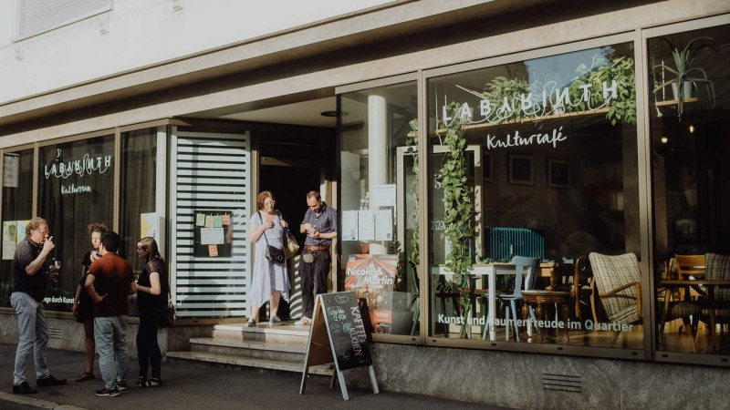 Menschen unterhalten sich vor dem LABYRINTH Kulturzentrum & -café. Die Fenster sind mit Pflanzen dekoriert, und ein Schild steht vor dem Eingang., © Christoph Steinweg Menschen unterhalten sich vor dem LABYRINTH Kulturzentrum & -café. Die Fenster sind mit Pflanzen dekoriert, und ein Schild steht vor dem Eingang., © Christoph Steinweg