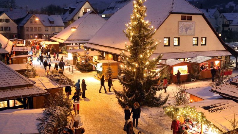 Verschneiter Weihnachtsmarkt in Metzingen mit beleuchtetem Baum und Ständen. Menschen schlendern durch die festliche Atmosphäre auf dem Kelternplatz., © Stadt Metzingen Verschneiter Weihnachtsmarkt in Metzingen mit beleuchtetem Baum und Ständen. Menschen schlendern durch die festliche Atmosphäre auf dem Kelternplatz., © Stadt Metzingen