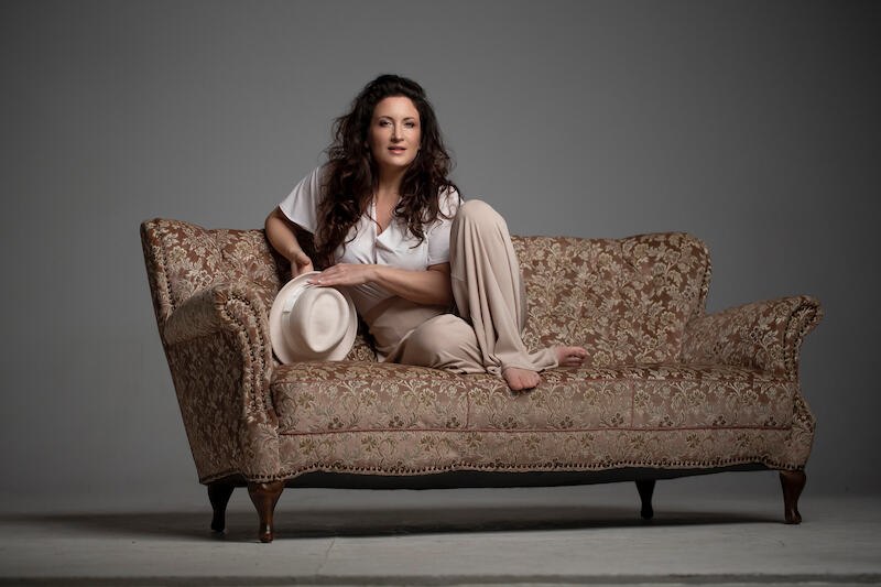 Eine Frau sitzt entspannt auf einem antiken Sofa, h&auml;lt einen Hut in der Hand und tr&auml;gt legere Kleidung. Der Hintergrund ist neutral gehalten., &copy; Sch&ouml;nblick gemeinn&uuml;tzige GmbH