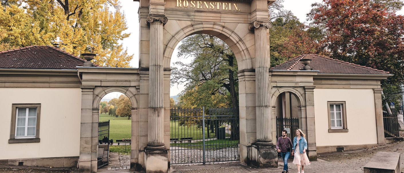 Eingangstor zum Rosensteinpark mit der Aufschrift 'Rosenstein'. Zwei Personen spazieren vorbei, umgeben von herbstlichen Bäumen., © SMG, Christoph Düpper