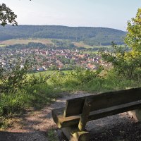 Eine Holzbank bietet einen malerischen Blick auf das Dorf Gündelbach, umgeben von grünen Feldern und bewaldeten Hügeln., © Land der 1000 Hügel - Kraichgau-Stromberg Eine Holzbank bietet einen malerischen Blick auf das Dorf Gündelbach, umgeben von grünen Feldern und bewaldeten Hügeln., © Land der 1000 Hügel - Kraichgau-Stromberg