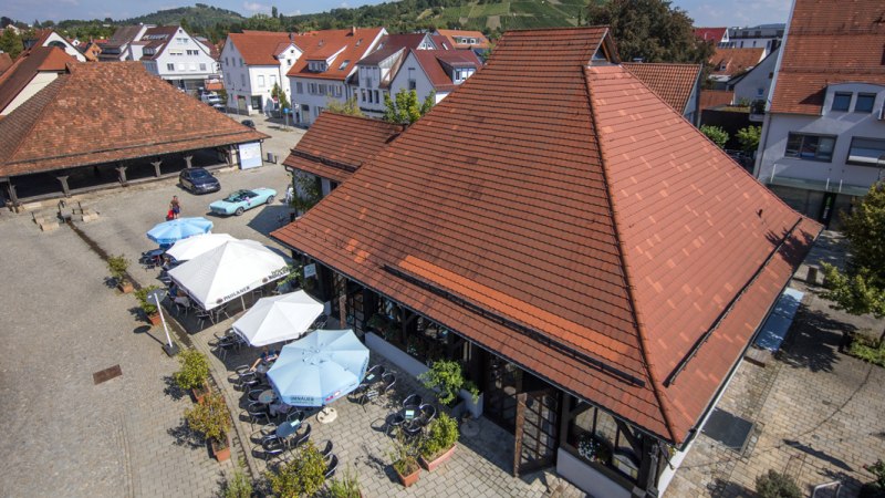Luftaufnahme der Sieben Keltern in Metzingen. Rote Ziegeldächer, Sonnenschirme und Tische auf einem gepflasterten Platz. Umgeben von Gebäuden und grünen Hügeln., © Stuttgart-Marketing GmbH Luftaufnahme der Sieben Keltern in Metzingen. Rote Ziegeldächer, Sonnenschirme und Tische auf einem gepflasterten Platz. Umgeben von Gebäuden und grünen Hügeln., © Stuttgart-Marketing GmbH