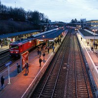 Bahnhof Backnang bei Dämmerung. Ein roter Zug steht am Gleis, Menschen warten auf den Bahnsteigen. Beleuchtung und Schienen sind sichtbar., © Alex Becher