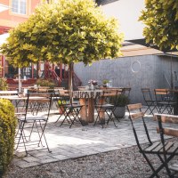 Ein leerer Café-Außenbereich mit Holztischen und -stühlen, umgeben von grünen Bäumen und Pflanzen, auf einem gepflasterten Boden., © © N. Trautmann