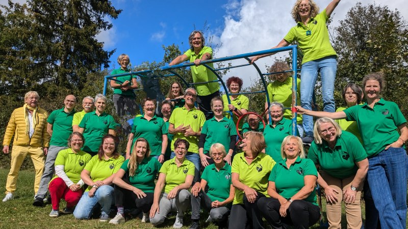 Gruppe in gr&uuml;nen T-Shirts posiert fr&ouml;hlich im Freien vor einem Kletterger&uuml;st. Einige stehen oben, andere sitzen oder stehen davor. Sonniges Wetter, blauer Himmel., &copy; Voice Factory
