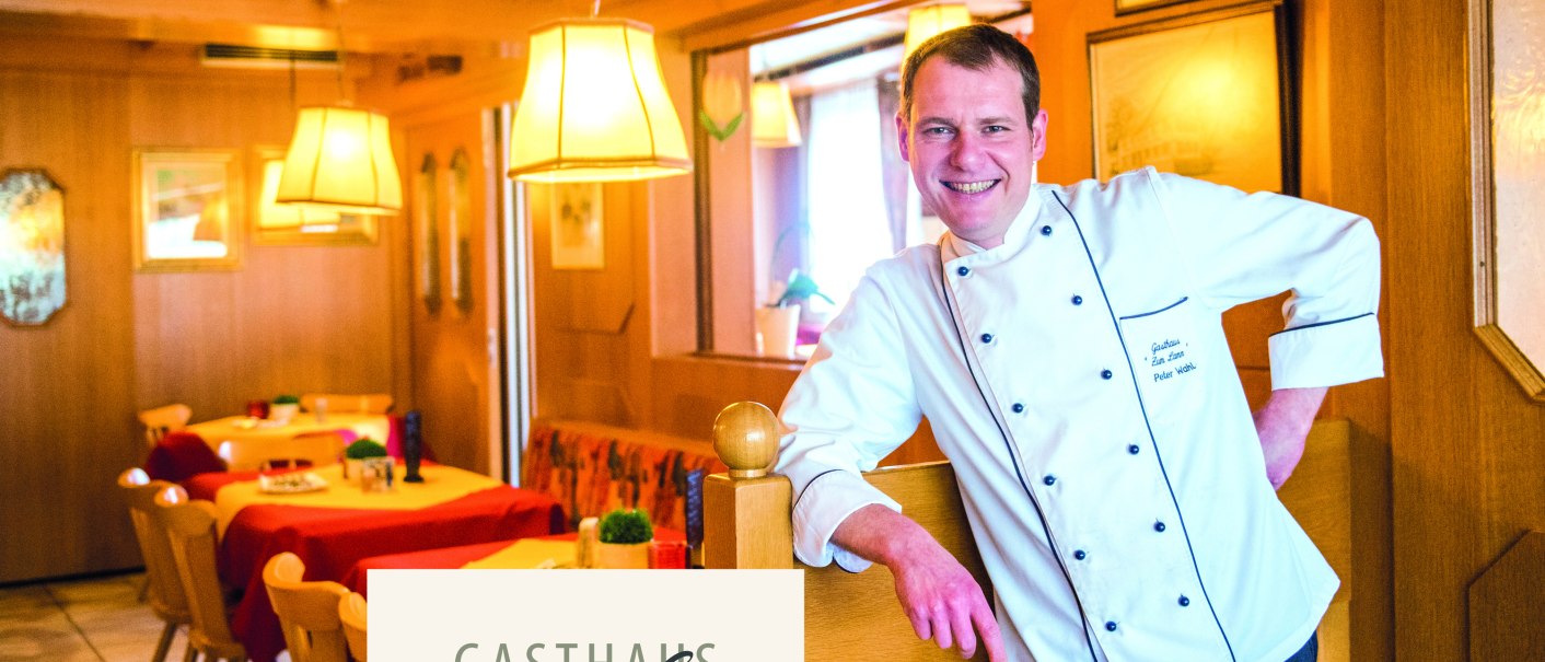 Ein Koch in weißer Uniform steht lächelnd in einem gemütlichen Gasthaus mit Holzmöbeln und roten Tischdecken., © Gasthaus Lamm Schornbach, Fotografie Nicole Schielberg Ein Koch in weißer Uniform steht lächelnd in einem gemütlichen Gasthaus mit Holzmöbeln und roten Tischdecken., © Gasthaus Lamm Schornbach, Fotografie Nicole Schielberg