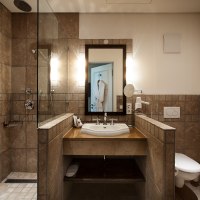 Modernes Badezimmer mit begehbarer Dusche, Waschbecken und Toilette. Die Wände sind mit braunen Fliesen verkleidet, ein Spiegel hängt über dem Waschbecken., © TOMAS