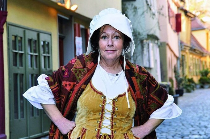 Frau in mittelalterlicher Kleidung mit Haube und Umhang steht auf einer gepflasterten Straße vor historischen Gebäuden., © Esslinger Stadtmarketing &amp; Tourismus GmbH