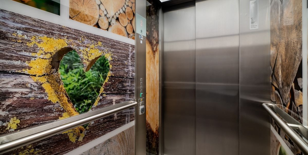 Ein Fahrstuhl mit Holzmuster und Herzmotiven an den Wänden. Die Türen sind aus Metall, und es gibt Handläufe an den Seiten., © Novum Hospitality Ein Fahrstuhl mit Holzmuster und Herzmotiven an den Wänden. Die Türen sind aus Metall, und es gibt Handläufe an den Seiten., © Novum Hospitality