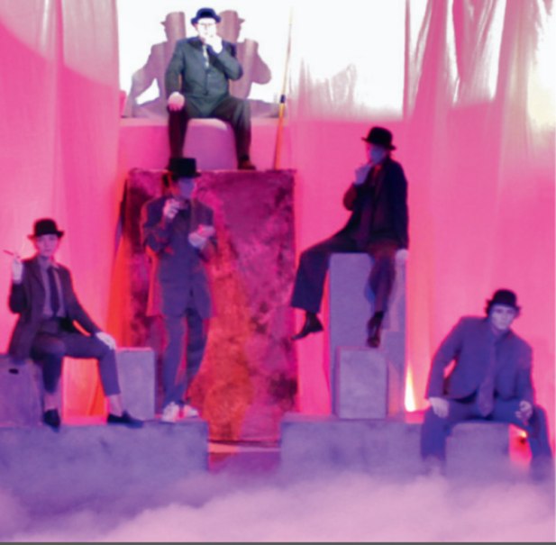 Fünf Personen in Anzügen und Hüten sitzen auf Podesten in rosa Licht. Die Szene wirkt theatralisch und mystisch., © Theaterschule an der Gmünder VHS