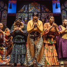 Eine Gospelgruppe singt auf der Bühne in bunten, traditionellen Gewändern vor einem dekorativen Hintergrund., © links im Bild
