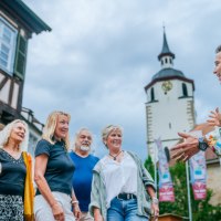 Eine Gruppe von Menschen lauscht einem Stadtf&uuml;hrer vor einem Fachwerkhaus und einem Turm mit Uhr. Der Himmel ist bew&ouml;lkt., &copy; Stadt Waldenbuch