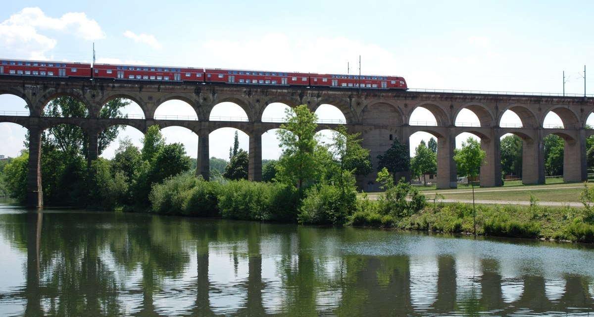 Ein roter Zug f&auml;hrt &uuml;ber das Enz-Viadukt, das sich &uuml;ber einen Fluss spannt. Der Himmel ist klar, und B&auml;ume s&auml;umen das Ufer., &copy; 3B-Tourismus-Team